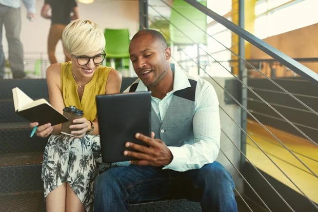 Two creative millenial small business owners working on social media strategy using a digital tablet while sitting in staircase-Flawless Inbound